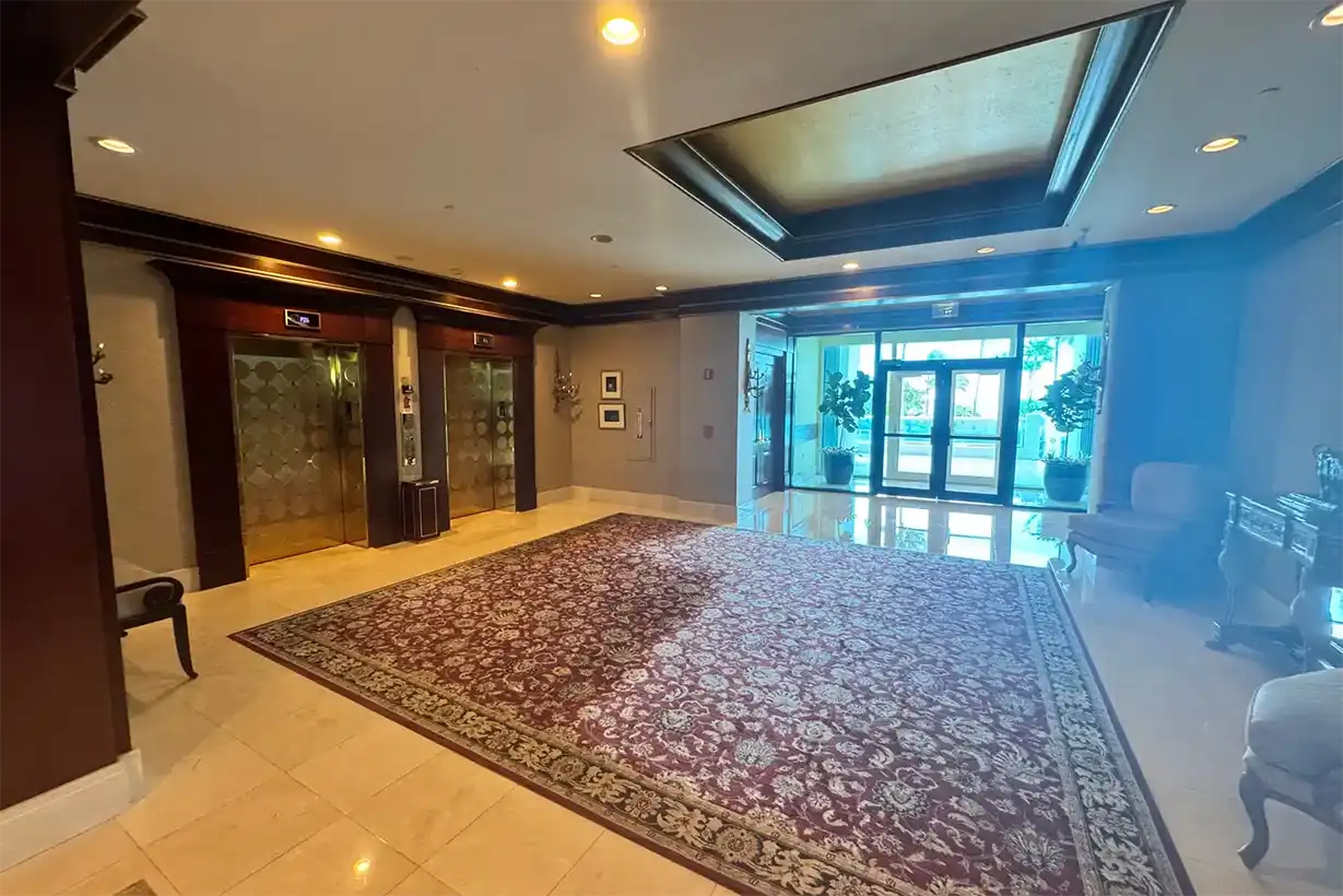 Hotel lobby entrance with metallic coffered ceiling detail, brass elevator doors, and view through glass doors to landscaped exterior