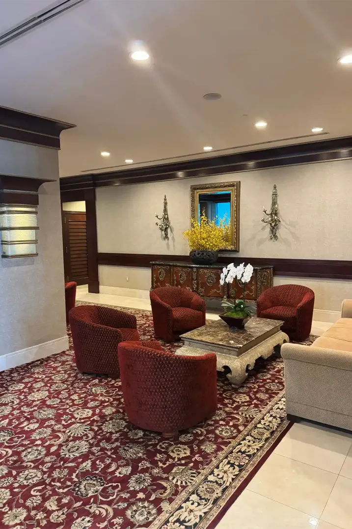 Traditional hotel seating lounge with ornate Persian carpet, burgundy upholstered barrel chairs, carved wood console, and dark millwork details